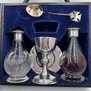 May include: A vintage silver communion set, comprising a chalice, two bottles, a spoon, and a cross-shaped piece, presented in a blue velvet-lined case with a black handle. The bottles are clear and dark, suitable for religious use.