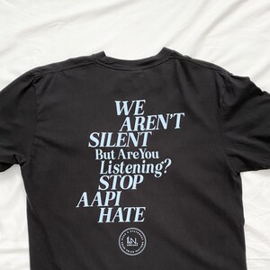 Puede incluir: Camiseta negra con un estampado gráfico azul claro que dice "WE AREN'T SILENT But Are You Listening? STOP AAPI HATE". El gráfico también incluye un pequeño círculo con el texto "LN DESIGNS MAKE A STATEMENT YOUR VOICE MATTERS".