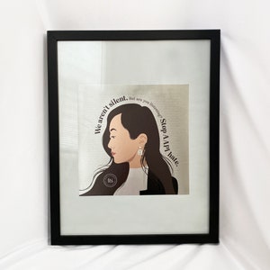 May include: A framed print with a black frame and a white background. The print features a digital illustration of a woman with brown hair and a black jacket. The text "We aren't silent. But are you listening? Stop AAPI hate." is written in a circle around the woman's head.
