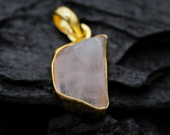 Può includere: Un ciondolo d'oro con una pietra preziosa rosa chiaro di forma irregolare. Il ciondolo è su uno sfondo nero.