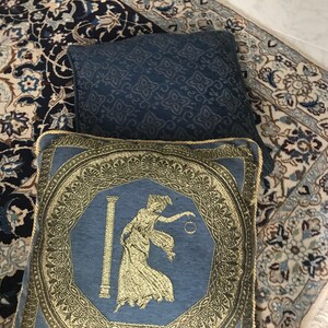 以下が含まれることがあります： 金色の刺繍が施された青いクッションが2つ。手前のクッションには、円形を持った女性が柱の横に立っている金色のデザインが施されています。