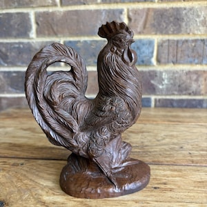 May include: A detailed, hand-carved wooden rooster figurine. The brown sculpture features intricate feather detailing, a prominent comb, and a curved tail. The rooster stands on a small, textured base, showcasing the craftsmanship of the carving.