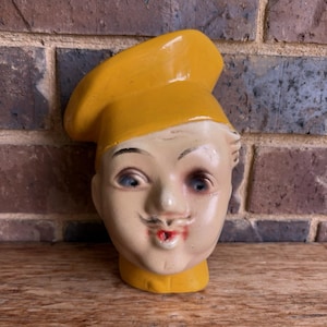 May include: A vintage ceramic chef head figurine with a yellow toque and collar. The face is painted with blue eyes, a small mustache, and red lips. The figurine is set against a brick wall and wooden surface.