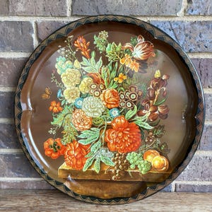 May include: Round, brown metal tray with a floral still life design. The tray features a detailed painting of colorful flowers, fruit, and butterflies. The rim has a decorative black and brown scalloped edge. A vintage decorative piece.