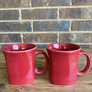 Puede incluir: Dos tazas rojas de cerámica con asa curva y borde ligeramente acampanado. Las tazas tienen estrías horizontales alrededor del cuerpo y se apoyan sobre una superficie de madera. El fondo es una pared de ladrillos.