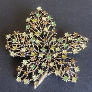 Puede incluir: Un broche de hoja de color dorado con un diseño de alambre detallado y una variedad de piedras de imitación verdes, marrones y transparentes.