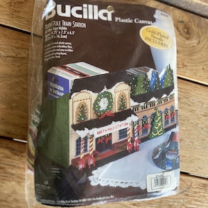 May include: A Bucilla plastic canvas kit for a North Pole Train Station. The kit includes a gold-plated needle and embroidery floss. The finished piece measures approximately 14.25" x 7.5" x 6.5". The packaging shows a completed cross-stitch design of a festive train station.