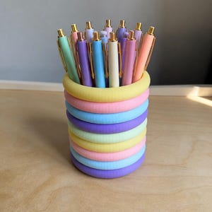 May include: A pen holder filled with pastel-coloured pens. The holder is made of stacked, rounded rings in shades of yellow, pink, blue, and purple. The pens have gold accents and come in various pastel colours, including green, purple, and pink.