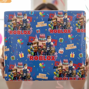 May include: A blue gift box wrapped in Roblox-themed birthday paper. The paper features Roblox characters, birthday cakes, gift boxes, and balloons. The words "Happy Birthday" and "Roblox" are also printed on the wrapping paper.