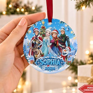 May include: A round ceramic Christmas ornament featuring a colorful illustration of characters from Frozen, including Elsa, Anna, Kristoff, Sven, and Olaf. The design includes the name Sophia and the year 2026 in blue text, suspended by a red ribbon.