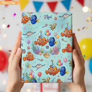 May include: A rectangular gift wrap with a repeating pattern of animated fish characters, party hats, balloons, and marine life on a light blue background. The wrap is held by two hands, with a festive party backdrop.