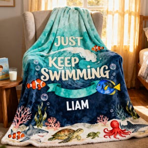 May include: A soft plush throw blanket featuring an ocean theme with a large wave, bubbles, and the text JUST KEEP SWIMMING and LIAM. The design includes a clownfish, a blue tang, a shark, a sea turtle, and a red octopus on a blue background.