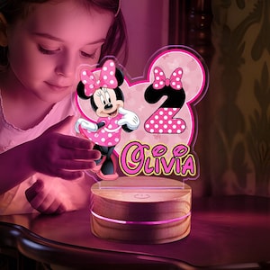 May include: A Minnie Mouse-themed night light with a pink and white polka dot design. The light features the number 2 and the name "Olivia" in a cursive font. The base is made of wood and emits a soft pink glow.