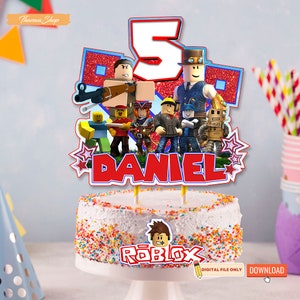 May include: A pink cake with colorful sprinkles and a red, blue, and white cake topper featuring Roblox characters, the number 5, and the name "DANIEL". The cake topper also includes the word "ROBLOX" in red letters.