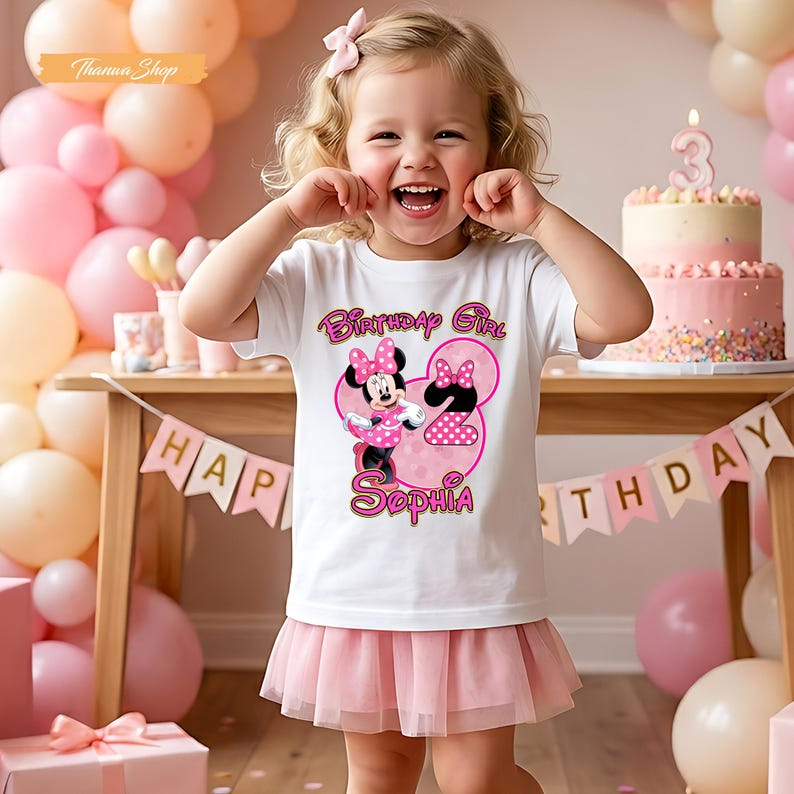 May include: A white short-sleeved t-shirt featuring a graphic of Minnie Mouse, the text Birthday Girl, the number 2, and the name Sophia in pink and black. The shirt is paired with a light pink tulle skirt, creating a festive birthday outfit.