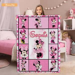 May include: Pink and white Minnie Mouse blanket featuring multiple images of Minnie Mouse in various poses. The blanket has a grid design with the name "Sophia" in the center. The blanket is held by a person.
