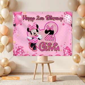 May include: A pink birthday banner featuring Minnie Mouse, the text "Happy 2nd Birthday Olivia", and polka dot accents. Balloons in shades of pink, gold, and white decorate the background. A small table holds a cake and a gift box.