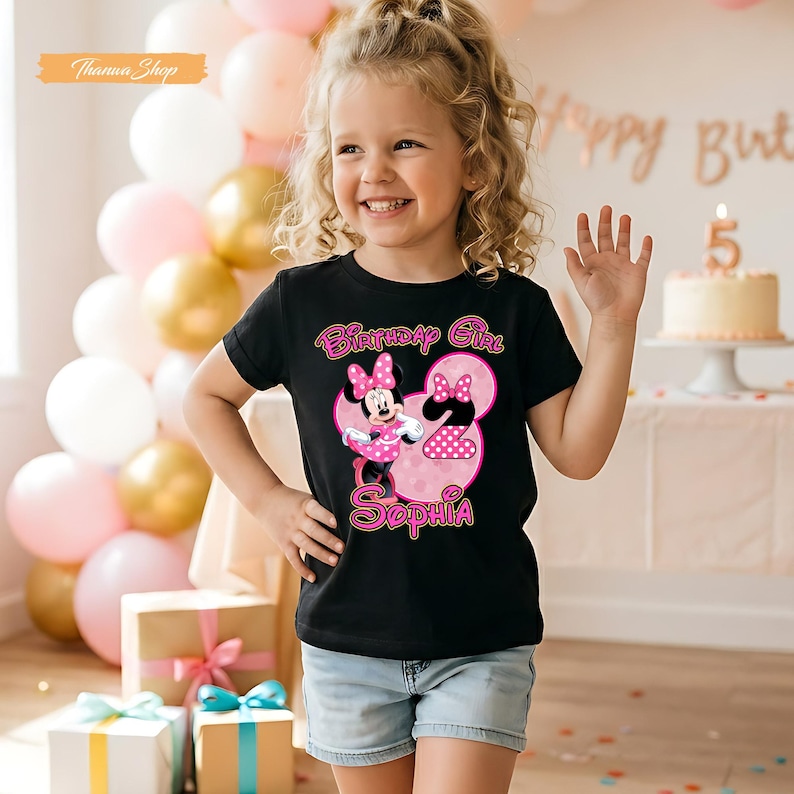 May include: A black short-sleeved t-shirt featuring a graphic of Minnie Mouse, a large pink number two, and the name Sophia in pink lettering. The text Birthday Girl is printed above the character. The design includes pink polka dot accents.