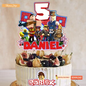 May include: A red, white, and blue cake topper featuring a group of Roblox characters, the number 5, and the name "DANIEL". The cake topper is on top of a white cake decorated with fruit and berries. The cake is on a wooden surface. The text "Roblox" is printed on the cake.