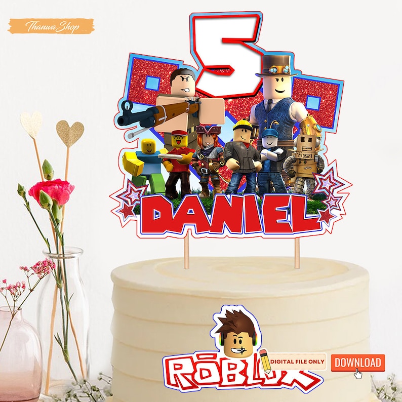 May include: A red, white, and blue Roblox cake topper with a number 5, the word "DANIEL", and a group of Roblox characters. The cake topper is on a white cake with the word "ROBLOX" and a Roblox character on the front.