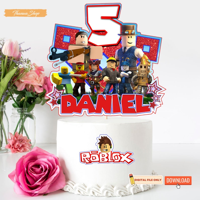 May include: A white cake with a red, blue, and white Roblox cake topper featuring a group of Roblox characters, the number 5, and the name "DANIEL". The cake topper is on a white cake with the word "ROBLOX" written in red and blue on the front. The cake is on a white surface with a pink rose in the foreground.