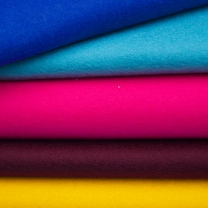 Może przedstawiać: Stosik pięciu kwadratowych kawałków filcu w różnych kolorach: niebieskim, jasnoniebieskim, różowym, bordowym i żółtym.