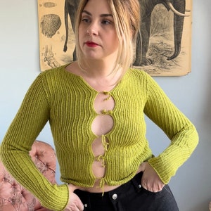 Könnte beinhalten: Ein limegrüner Strickcardigan mit einem Ausschnitt-Design und einem Bindedetail an der Vorderseite. Der Cardigan ist kurz und hat lange Ärmel.