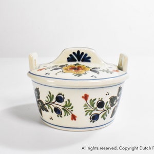 Puede incluir: Un salero de cerámica color crema con tapa, adornado con motivos florales y de bayas en azul, amarillo y rojo. El salero tiene un asa en la tapa y dos asas pequeñas en los laterales. El diseño recuerda a la cerámica de Delft holandesa.