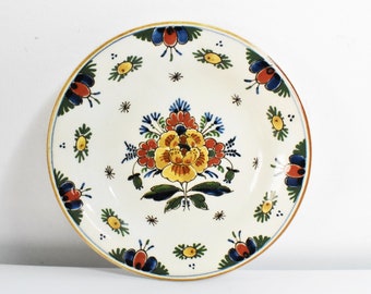 Könnte beinhalten: Ein weißer Keramikteller mit handgemaltem Blumendesign. Der Teller zeigt eine zentrale gelbe Blume, umgeben von roten, blauen und grünen Akzenten. Der Rand hat einen goldenen Rand. Ein dekoratives Stück für das Zuhause.