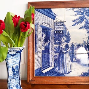 May include: A blue and white Delft tile artwork in a wooden frame depicts a Dutch scene with figures and text. A blue and white vase with a landscape design holds red tulips, adding a pop of color to the scene.