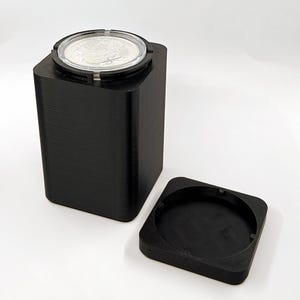 May include: A black coin holder with a clear top, displaying a silver coin. The holder is open, with the lid separate, showing the coin. The holder is made of a textured material.