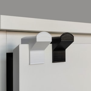 Op de afbeelding: Twee haken, een wit en een zwart, zijn bevestigd aan een wit oppervlak. De haken zijn gemaakt van plastic en hebben een afgeronde bovenkant.