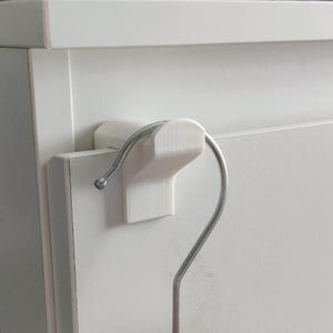 May include: A white, 3D-printed hanger hook attached to a white cabinet door. The hook is designed to hold a metal clothes hanger. The design is simple and functional, providing a space-saving storage solution.