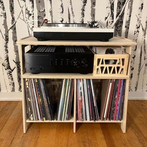 Peut inclure: Un meuble pour platine vinyle en bois clair avec une platine noire et blanche sur le dessus. Un amplificateur stéréo noir est en dessous, avec une caisse en bois sur le côté. L'étagère inférieure contient une collection de disques vinyles.