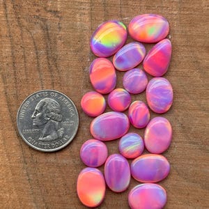 May include: A collection of seventeen smooth, oval-shaped cabochons in shades of pink, purple, and orange with iridescent finishes, displayed next to a United States quarter dollar coin for scale to show their relative size.