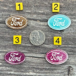 May include: Four oval-shaped Ford emblems with different designs and colors. One has a leopard print background, another is light blue, and two have pink patterns. A US nickel coin is also present.