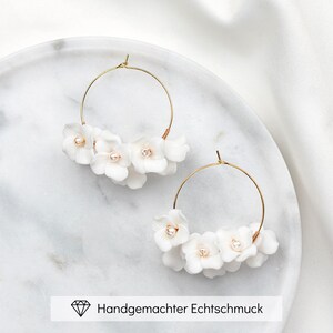 Könnte beinhalten: Ein Paar goldene Creolen mit weißen Blumenclustern und kleinen Perlenakzenten. Die Ohrringe werden auf einer weißen Marmoroberfläche präsentiert. Der Text "Handgemachter Echtschmuck" befindet sich unten.