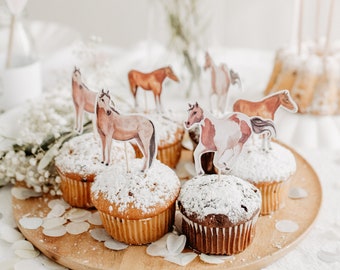Puede incluir: Un plato de madera con seis cupcakes decorados con azúcar en polvo y adornos de caballos. Los cupcakes están sobre un mantel blanco con confeti blanco.