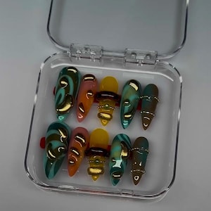 Op de afbeelding: Een set van tien kunstnagels in een doorzichtig plastic doosje. De amandelvormige nagels hebben diverse ontwerpen met blauwgroene wervelingen, effen geel en oranje gradiënten, allemaal voorzien van verhoogde gouden 3D-versieringen.