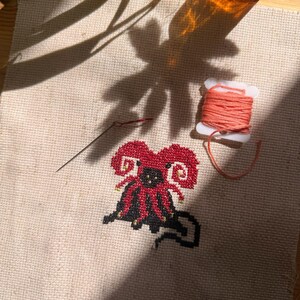 May include: A cross-stitch project on beige Aida fabric featuring a red and black creature with curled horns. A silver needle lies to the left of the design, and a white plastic spool wrapped with coral-colored embroidery floss sits to the right.