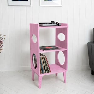 May include: A pink wooden record player stand with three shelves and oval cutouts. A record player sits on top, with a vinyl record and several album covers on the shelves. The stand is in a room with a black leather armchair and a vase of flowers.