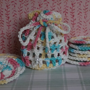 May include: A crocheted drawstring bag and matching coasters in white, yellow, pink, and blue. The bag has a lattice design and is tied with a pink and white cord. The coasters are stacked and scattered, showcasing the same color scheme.