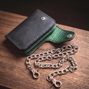 Op de afbeelding: Een zwarte leren portemonnee met een groene binnenkant en een zilveren ketting. De portemonnee is rechthoekig en heeft een druksluiting. De ketting heeft twee sluitingen. De portemonnee en de ketting liggen op een houten oppervlak.
