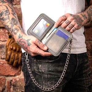 May include: Gray leather wallet with a silver chain, open to reveal credit cards and cash. The wallet has a clear window for a card. The chain is attached to the wallet with a small metal loop. The wallet is held by a person wearing a white shirt and black jeans.