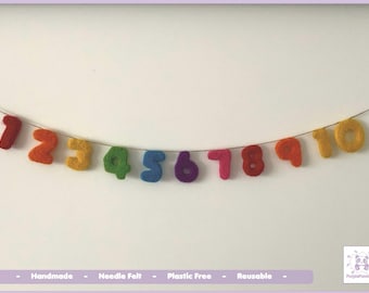 May include: A garland featuring ten individual needle felted numbers from one to ten, each in a distinct vibrant color including red, orange, yellow, green, blue, purple, and pink, all strung together on a thin, light-colored cord.