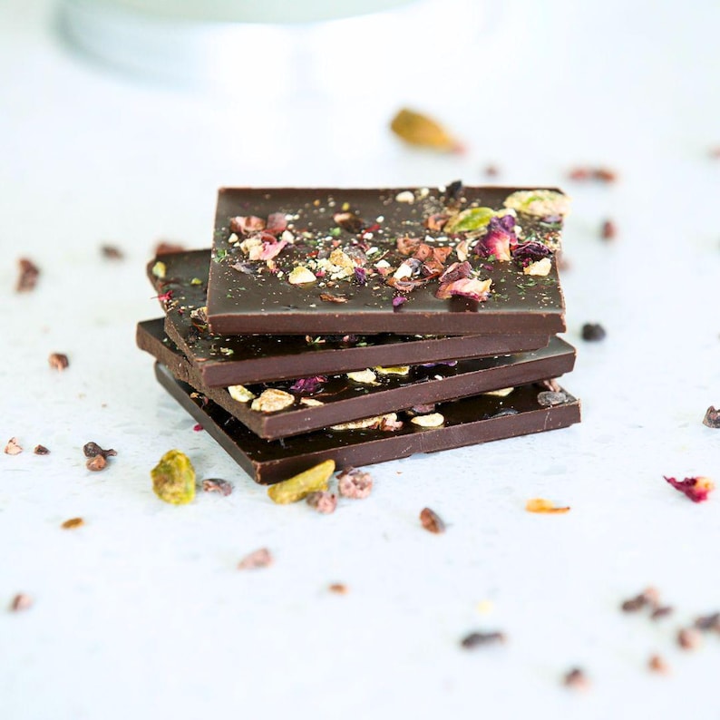 May include: A stack of four dark chocolate squares, each topped with colorful pistachios, dried rose petals, and cocoa nibs. The chocolate is a rich brown color, and the toppings add texture and visual interest. Scattered ingredients surround the stack.