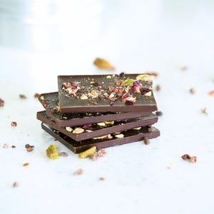 May include: A stack of four dark chocolate bars, each topped with colorful sprinkles of pistachios, dried rose petals, and other ingredients. The chocolate bars are arranged on a white surface, with scattered toppings.