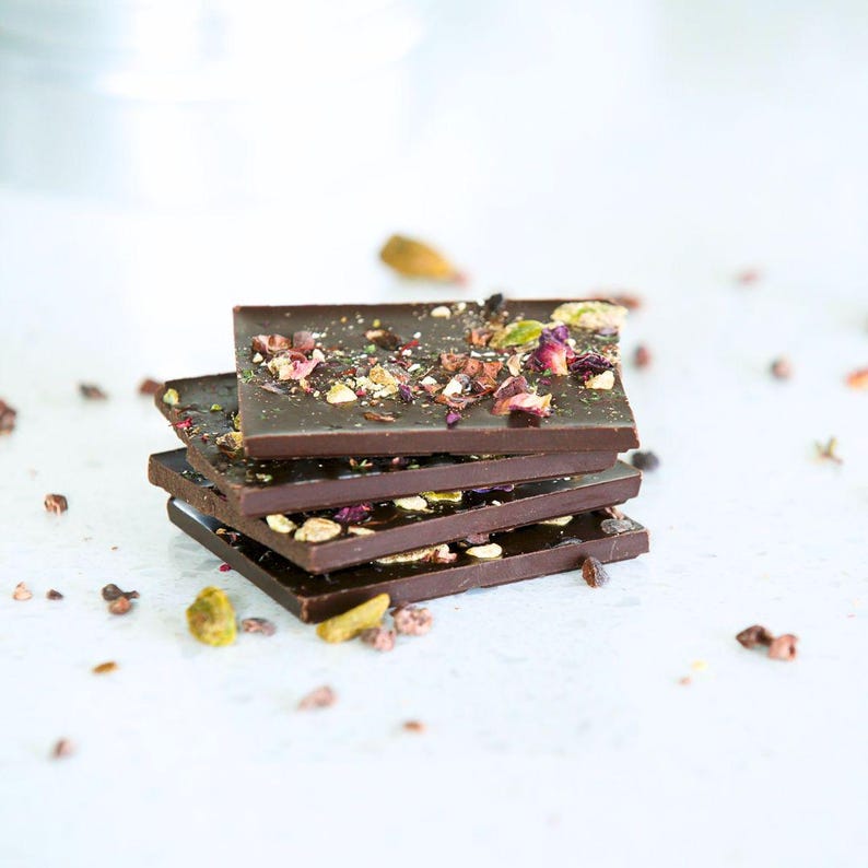 May include: A stack of four dark chocolate bars, each topped with colorful sprinkles of pistachios, dried rose petals, and other ingredients. The chocolate bars are arranged on a white surface, with scattered toppings.