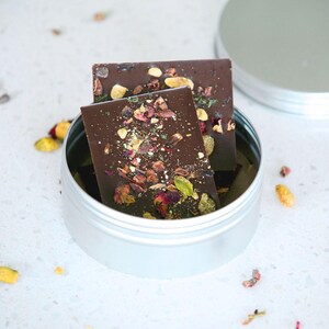 May include: Two dark chocolate bars with visible toppings, including pistachios and dried flowers, rest inside a silver tin. The tin's lid is partially open, revealing the contents. The chocolate bars are rectangular and appear to be artisan-made.