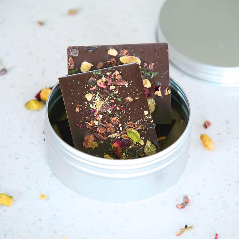 May include: Two dark chocolate bars with visible toppings, including pistachios and dried flowers, rest inside a silver tin. The tin's lid is partially open, revealing the contents. The chocolate bars are rectangular and appear to be artisan-made.
